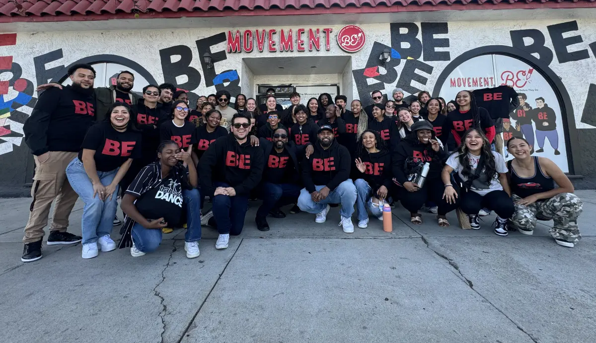 A large group of young people and staff in black BE hoodies pose together outside the mural-covered Movement BE center.