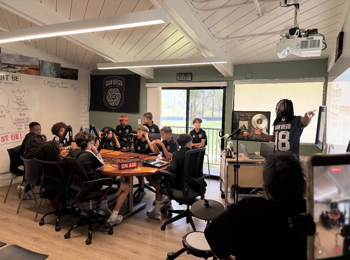 A mentor in a number 18 jersey leads a podcast session as students in headphones sit around a studio table of microphones.