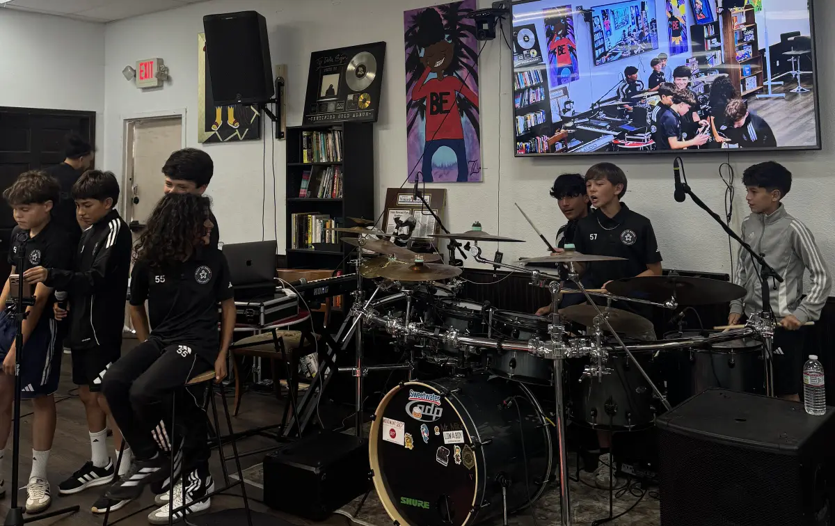 Young musicians in matching academy kits perform on drums and microphones in the Movement BE studio while a screen shows them live.