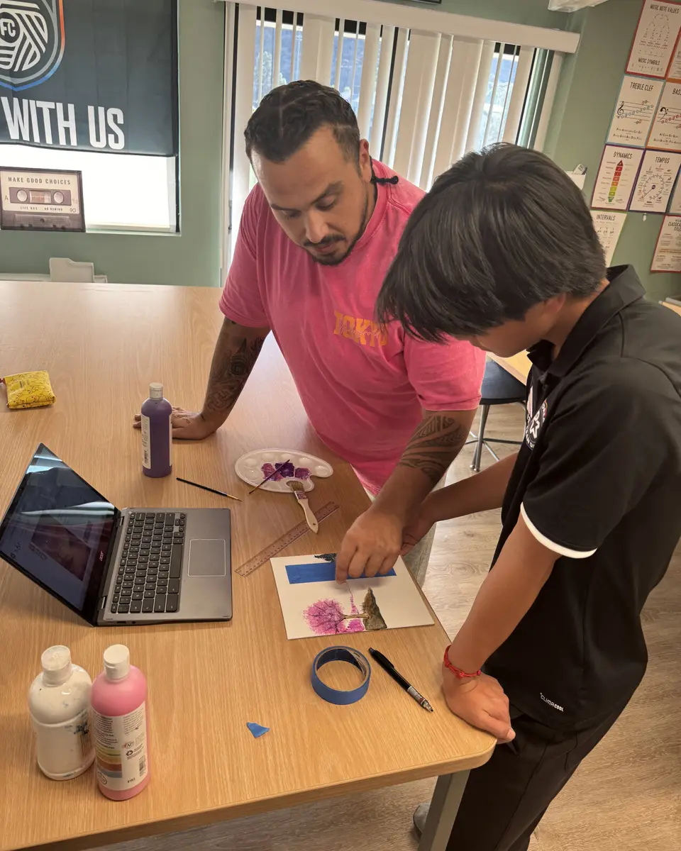 A mentor leans over a table to guide a student working on a painting, with paints and brushes beside them.
